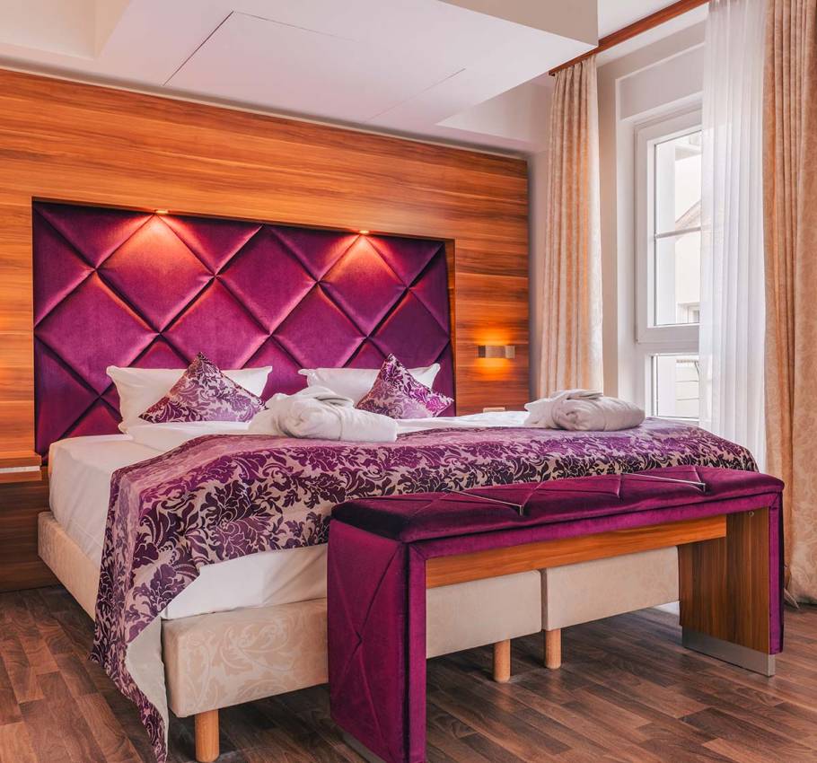 Doppelbett Suite in der Ferienwohnung in Binz auf Rügen mit violettem Kopfteil und eleganter Bettbank vor hellem Fenster.