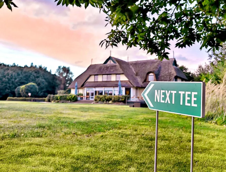 Exterior view of the golf center at sunset. In the foreground, a sign points to the next tee.