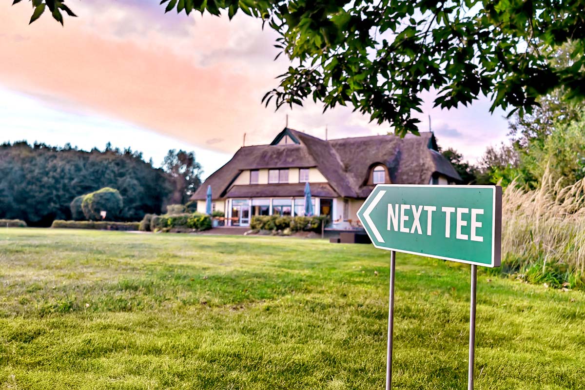 Außenansicht des Golfcentrums bei Sonnenuntergang. Im Vordergrund verweist ein Schild auf den nächsten Abschlag.